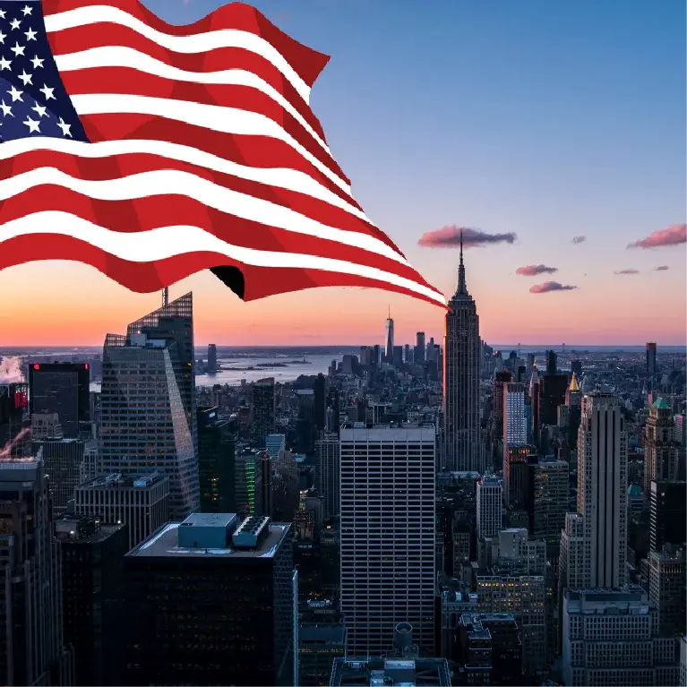Image of the American flag waving with the New York City skyline, including the Empire State Building, at sunset.